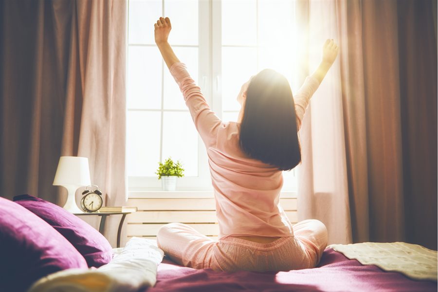 Egy nő az ágyán ülve nyújtózik a napfényes ablak előtt
